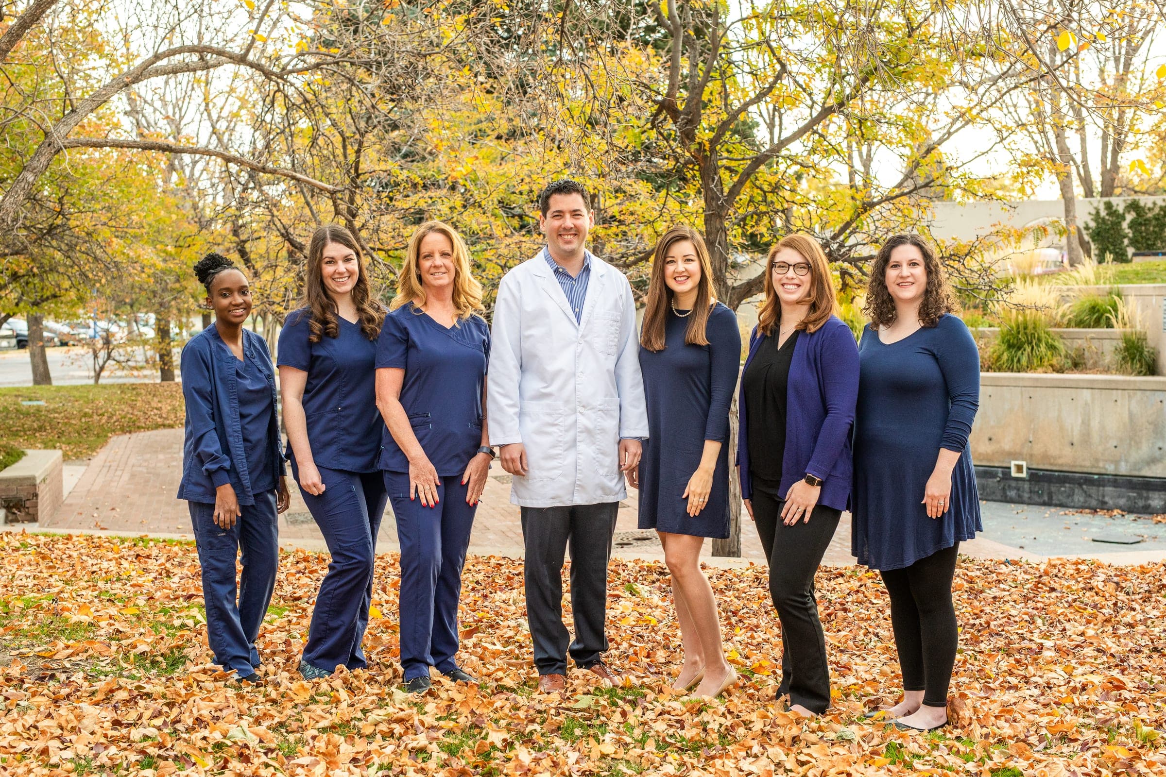 Dr. Tom Gutberg in a white coat with six Blue Sky Smiles team members in navy, standing in autumn leaves.